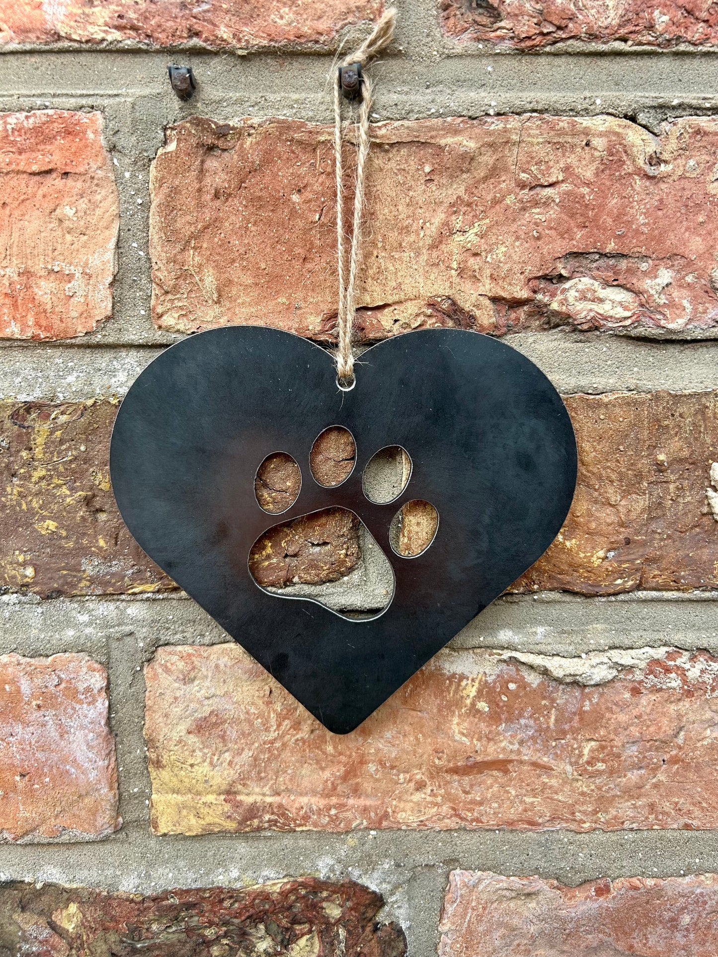 Steel Love Heart with Paw Print Cut Out hanging by a piece of string against a brick wall.