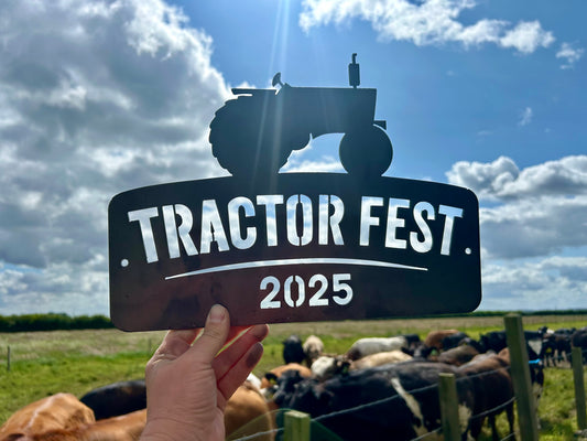 Hand holding a 'Tractor Fest 2025' sign with cows and a tractor in the background.