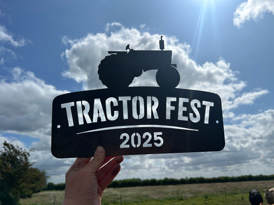 Hand holding a 'Tractor Fest 2025' sign against a cloudy sky