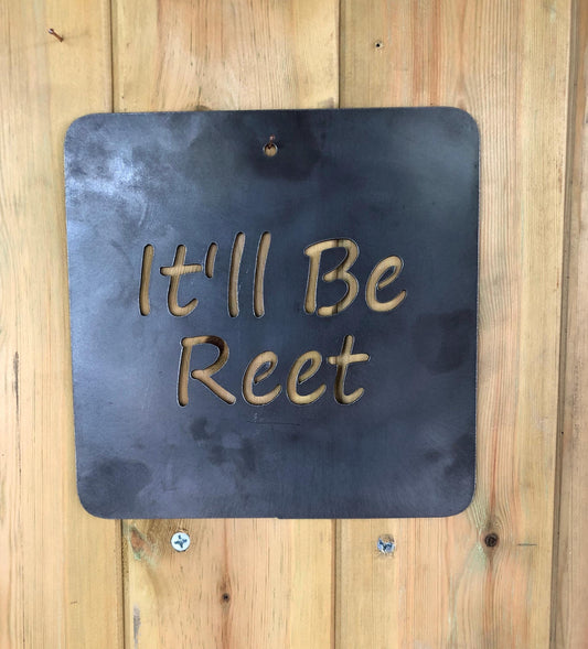 A steel wall sign with the phrase 'It'll Be Reet' written in a heart shape, displayed against a rustic wooden background.