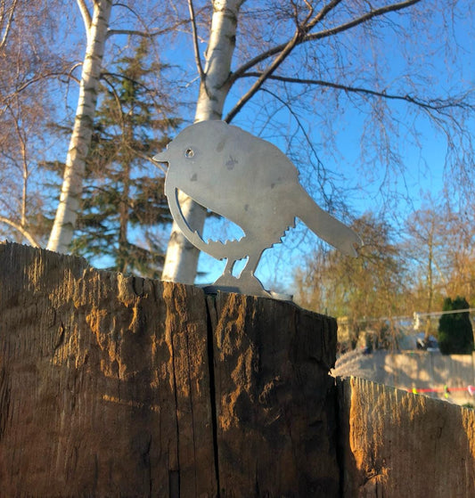 A metal fence topper in the shape of a rusty robin bird, placed on a wooden surface with foliage in the background.