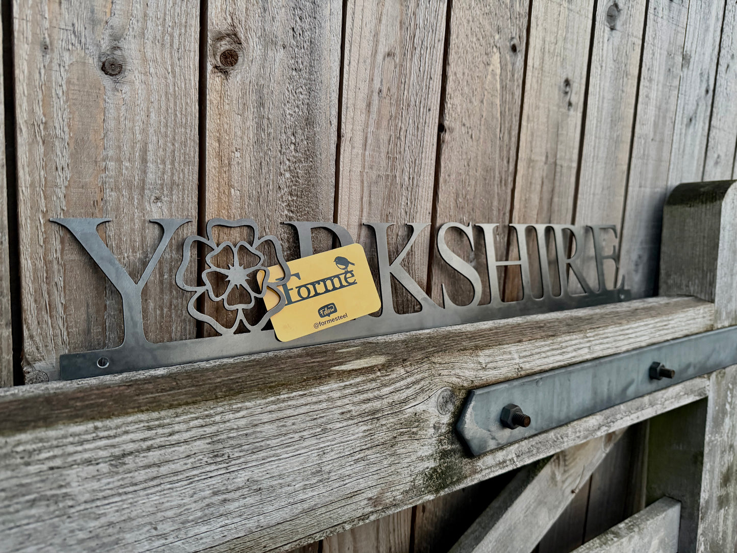 Hand holding a 'Yorkshire' sign with the 'o' replaced with a Yorkshire Rose, against a wooden background. Displaying a Forme Steel business card.