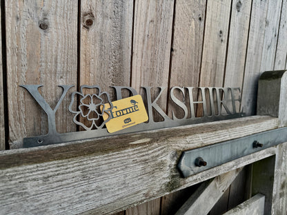 Hand holding a 'Yorkshire' sign with the 'o' replaced with a Yorkshire Rose, against a wooden background. Displaying a Forme Steel business card.