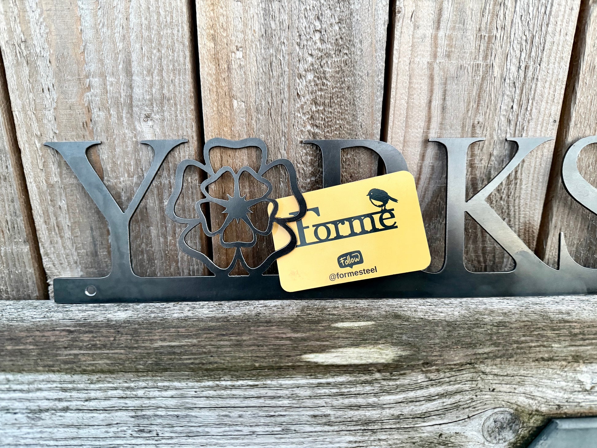 Hand holding a 'Yorkshire' sign with the 'o' replaced with a Yorkshire Rose, against a wooden background. Displaying a Forme Steel business card.