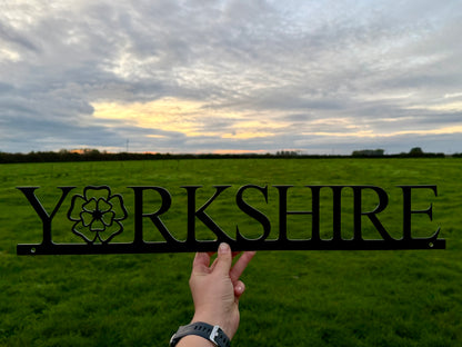 Hand holding a 'Yorkshire' sign with the 'o' replaced with a Yorkshire Rose, against a cloudy sky and field 