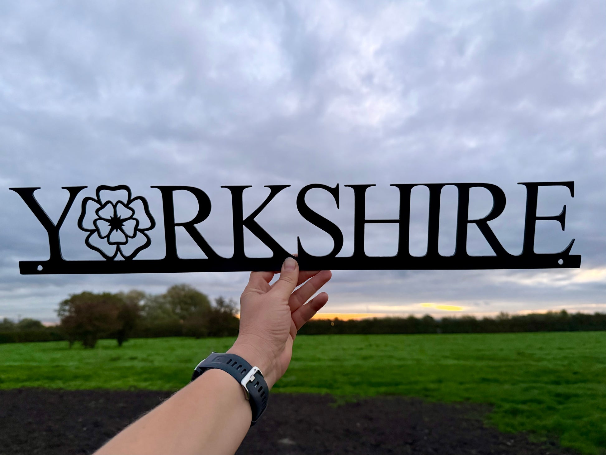 Hand holding a 'Yorkshire' sign with the 'o' replaced with a Yorkshire Rose, against a cloudy sky and field background