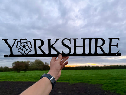 Hand holding a 'Yorkshire' sign with the 'o' replaced with a Yorkshire Rose, against a cloudy sky and field background