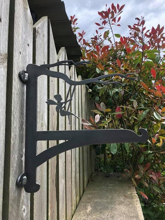 A metal bracket with a fairy on a swing design attached to a wooden fence, intended as use of a hanging basket bracket for hanging plants.